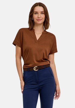 Femme portant un haut marron à manches courtes, un pantalon bleu et une ceinture foncée avec une boucle dorée, debout avec une main dans la poche.