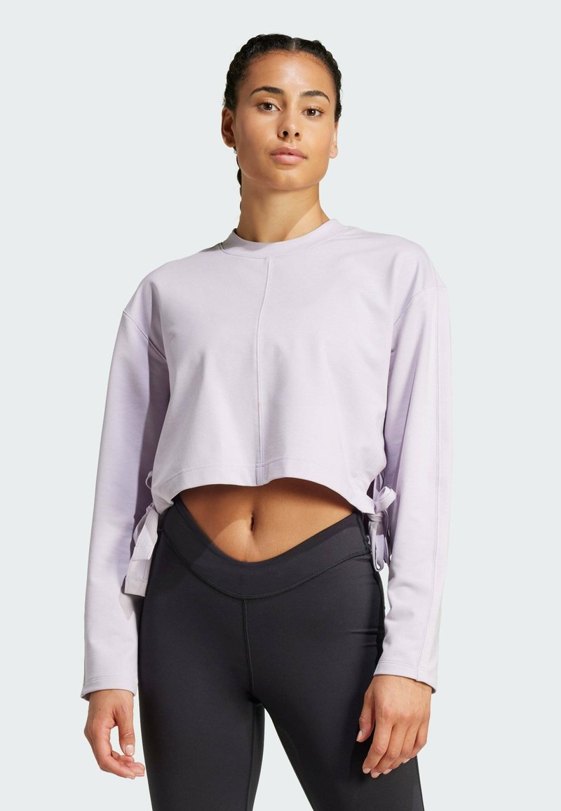 Korte langmouwtop van licht lavendel stof met een ronde halslijn en zijkoordjes, gecombineerd met zwarte high-waisted leggings.