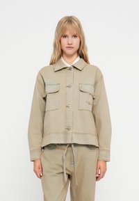 Beige, kort denimjakke med to frontlommer, sølvknapper og diskret s�øm. Har krage og snøring i kanten.