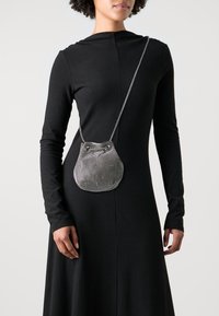 Borsa a tracolla in rete metallica con fondo arrotondato, chiusura a duplice occhio e una sottile tracolla a catena, indossata sopra un vestito nero a maniche lunghe.