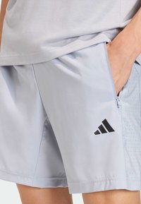Lyseblå atletiske shorts i et glatt stoff med svarte detaljer. Har en glidelåslomme og perforert mønster på den ene siden.