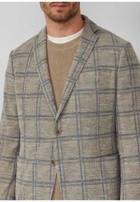 Braun kariertes Blazer aus strukturiertem Stoff mit blauen Gitterlinien, zwei Knöpfen und einem Revers über einem beigen Pullover.