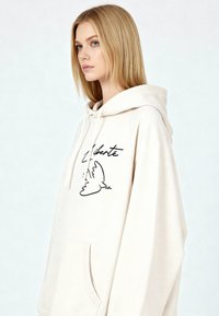 Felpa beige con tasca frontale, caratterizzata da una grafica nera di una colomba e la parola "Liberté". Tessuto morbido, vestibilità comoda, cappuccio con coulisse.