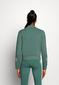 Sudadera deportiva verde con un alto cuello, cierre de cremallera y dobladillo acanalado. Presenta tres franjas blancas en cada manga. Textura suave.