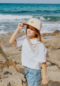 Cappellino da baseball beige con una toppa testurizzata, t-shirt bianca a maniche corte e jeans azzurri chiari, in piedi su rocce vicino all'oceano.