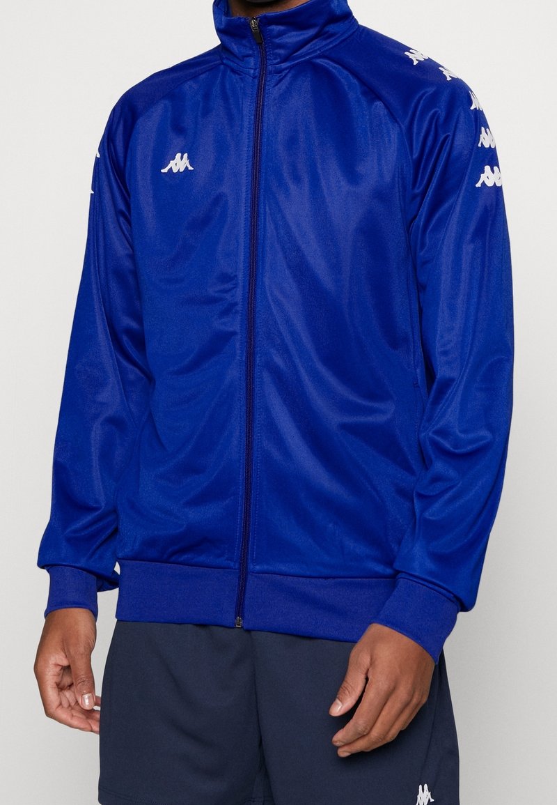 Blå sportjacka med hög krage, dragkedja och ribbade muddar. Har vita logodetaljer på axlarna. Tillverkad av slätt, lätt material.