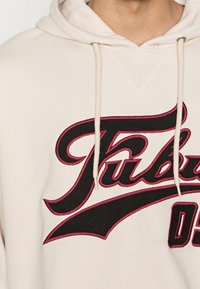 Beige hoodie met een voorkant v-hals, voorzien van gedurfde zwarte en rode geborduurde tekst en uniforme koorden met metalen uiteinden.