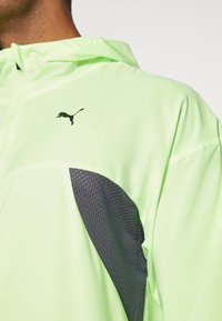 Veste de sport verte néon avec un panneau en maille noire respirante, dotée d'une capuche zippée et d'un petit logo noir sur la poitrine.