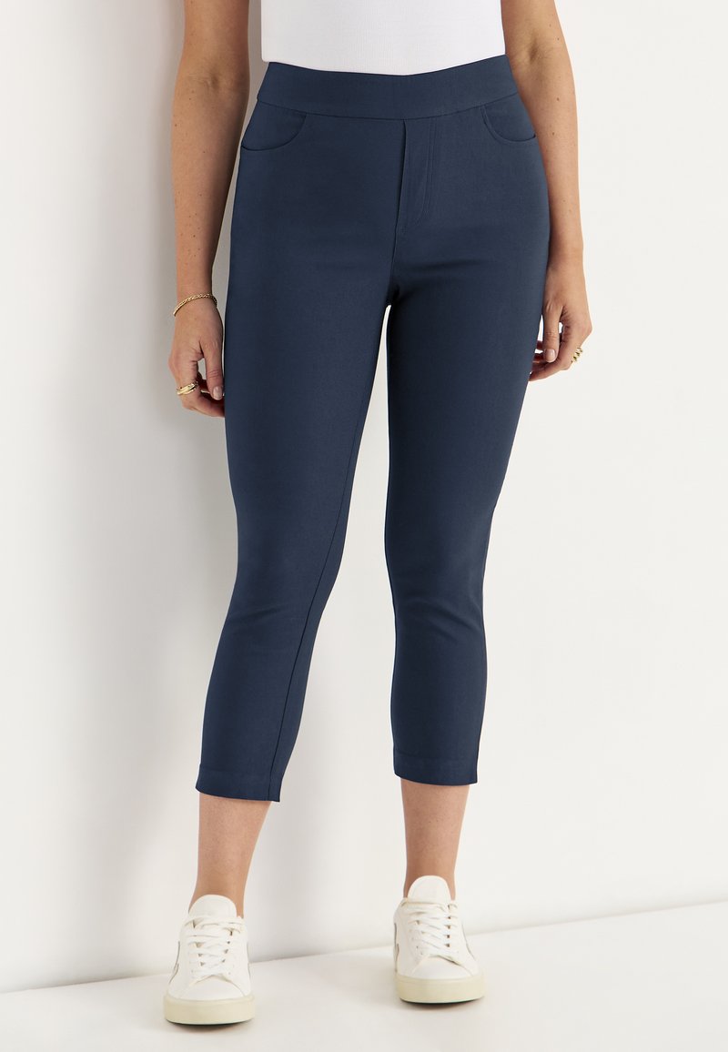 Frau trägt marineblaue Cropped-Hose und weiße Sneakers, steht vor einem einfarbigen hellen Hintergrund.
