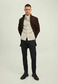 Casaco castanho sobre uma camisa bege de botão, combinado com jeans de denim escuro e botas pretas de atacador. Textura suave e corte ajustado.