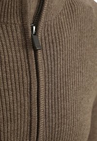 Bruine kleding met rits, gekenmerkt door ribbeltextuur en een zwarte ritstrekkertje. De stof lijkt zacht en warm, geschikt voor koelere weersomstandigheden.