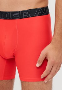 Boxer-shorts élastiques rouges avec une taille noire arborant "UNDER ARMOUR" en gris. Texture lisse et design ajusté, mettant en valeur la jambe.