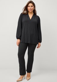 Blouse noire à manches longues avec un col en V et des poignets blousés, assortie à un pantalon texturé gris foncé et des chaussures à talons hauts nude.