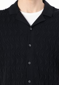 Cardigan noir en tricot texturé avec un motif en vagues, un col chemise et cinq boutons noirs, porté sur une chemise blanche à côtes.
