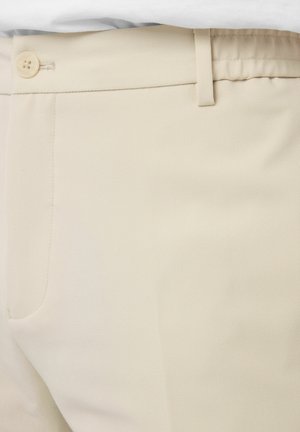 Gros plan sur un pantalon beige avec une ceinture boutonnée, passants pour ceinture et une poche latérale, le tout dans une texture de tissu lisse.