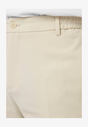 Gros plan sur un pantalon beige avec une ceinture boutonnée, passants pour ceinture et une poche latérale, le tout dans une texture de tissu lisse.