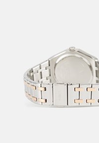 Bracelet de montre en métal argenté avec des accents en or rose, boîtier octogonal, finition polie et fermoir visible avec le nom de la marque gravé.