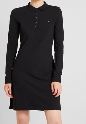 Schwarzes Polo-Kleid mit langen Ärmeln, einer fünf-Knopf-Leiste und kleinem Logo in Rot, Weiß und Blau auf der Brust, getragen von einer Person mit entspannt hängenden Händen.