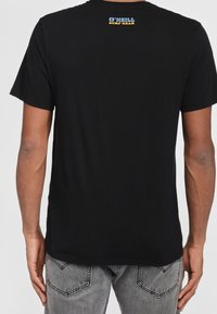 Camiseta de algodón negra de manga corta, con un logo rectangular en la parte superior de la espalda en amarillo y azul. La tela parece suave y sin patrón.