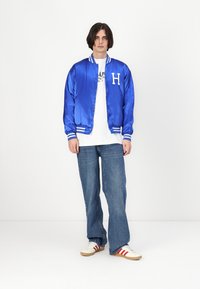 Jachetă bomber din satin albastru cu manșete și guler cu dungi albe, având un mare logo alb „H” pe partea stângă a pieptului, asortată cu blugi largi din denim și adidași.