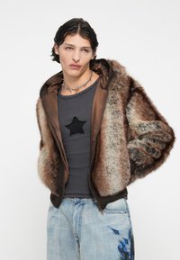 Jachetă din blană artificială maro cu fermoar în față, având glugă și manșete cu nervuri. Purtați-o peste un tricou gri cu un patch în formă de stea neagră și blugi de un albastru deschis.