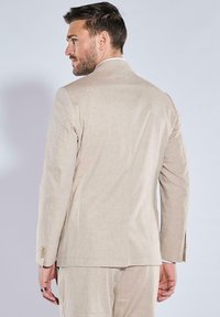 Hellbeige, maßgeschneiderte Sakko aus Leinen, mit einem Rückenschlitz, schmaler Passform und goldfarbenen Knöpfen an den Manschetten. Von hinten gezeigt.