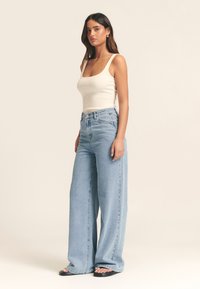 Gaiši krēmveida ribotais tops bez piedurknēm, kombinēts ar augsta vidukļa, platu kāju ziliem džinsiem. Džinsiem ir klasiska silueta un izbalināts mazgājums.