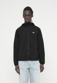 Μαύρο ελαφρύ μπουφάν με φερμουάρ και κουκούλα, με μικρό λογότυπο στο στήθος και ελαστικά μανίκια. Φορεμένο πάνω από ανοιχτόχρωμα μπλε τζιν.