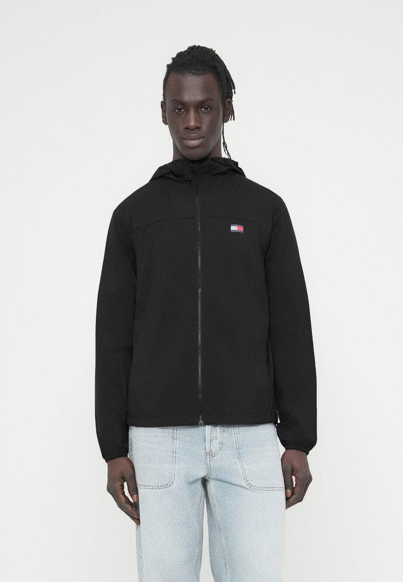 Μαύρο ελαφρύ μπουφάν με φερμουάρ και κουκούλα, με μικρό λογότυπο στο στήθος και ελαστικά μανίκια. Φορεμένο πάνω από ανοιχτόχρωμα μπλε τζιν.