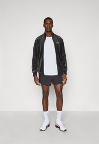 Γκρι βελούδινο μπουφάν με φερμουάρ, λευκό μπλουζάκι, σκούρες βερμούδες, λευκές κάλτσες με λογότυπο και αθλητικά παπούτσια με κόκκινες λεπτομέρειες, σε ουδέτερο φόντο.