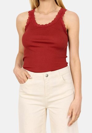 Vrouw draagt een mouwloze geribde rode top met kanten afwerking en hooggetailleerde ecru broek, hand in de zak op een neutrale achtergrond.