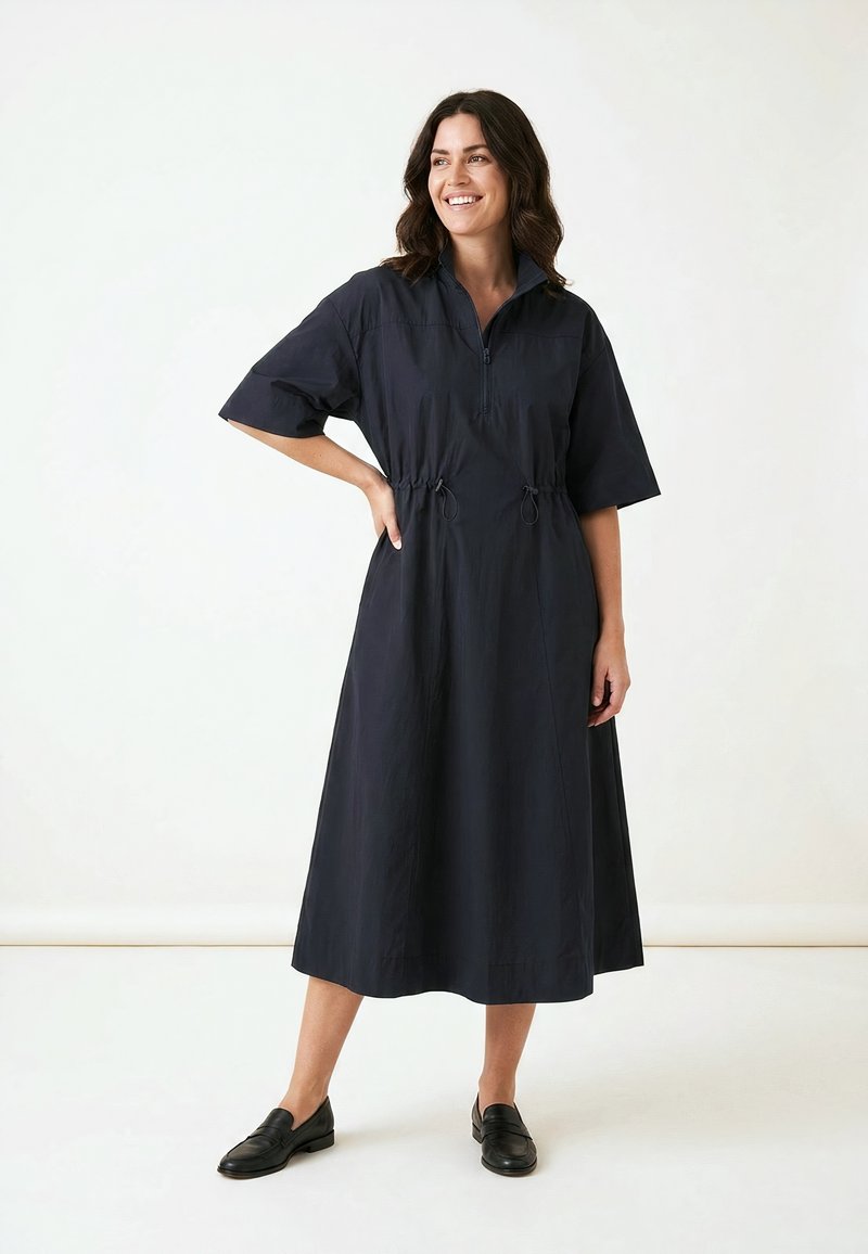 Femme debout souriante, portant une robe midi noire avec manches mi-longues, col zippé, taille cintrée et chaussures noires sans lacets sur fond blanc.