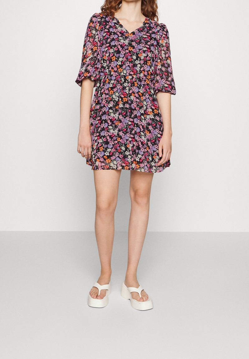 Femme portant une robe à manches courtes à imprimé floral en violet, rose, orange et noir, associée à des sandales compensées blanches.
