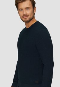 Pull en maille bleu marine avec une finition texturée, col rond, et poignets et ourlet côtelés. Présente une petite étiquette en tissu sur le côté.