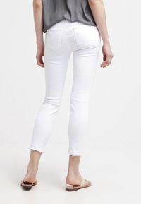 Jeans skinny de mezclilla blanca con una longitud recortada, dobladillo deshilachado y dos bolsillos traseros, combinados con sandalias deslizantes de un tono marrón claro.