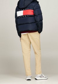 Marinblå pufferjacka med röd- och vit accentpanel, beige byxor och grå sneakers. Huva design med broderad logotyp på baksidan.