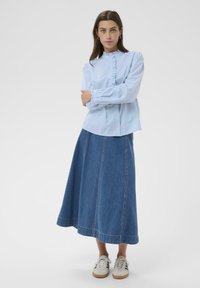 Chemise bleu clair à boutons avec des détails à volants, associée à une longue jupe en denim. Portée avec des baskets blanches. Tissu lisse et coupe décontractée.