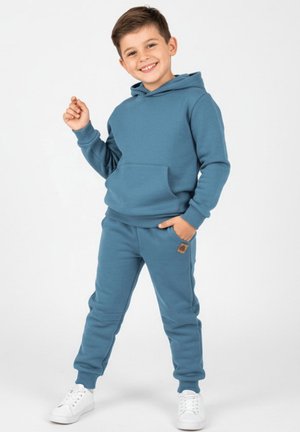 Niebieska bluza z kapturem i pasujące do niej spodnie dresowe wykonane z miękkiego materiału, z kieszenią kangurka, ribowanymi ściągaczami i małą naszywką z logo na spodniach.