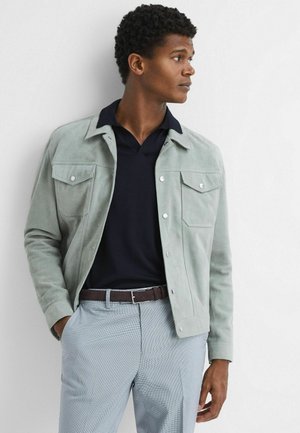 Hellgrüne Wildlederjacke mit zwei Brusttaschen, silbernen Knöpfen, kombiniert mit einem navyblauen Polo und grauen gemusterten Hosen. Glatte Textur überall.