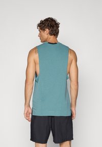 Turkisærmeløs tanktop med rund halsudskæring og åbne sider, lavet af blødt stof; båret med sorte sportsshorts.