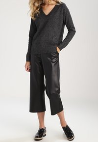 Dunkelgrauer V-Ausschnitt-Pullover mit strukturiertem Strick, kombiniert mit schwarzen, cropped Hosen aus Lederimitat. Das Model trägt schwarze Plateauschuhe.