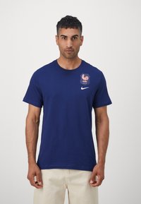 Tamsiai mėlynas medvilninis marškinėlis su šviesiai rožiniu gaidžio emblema ir baltu Nike logotipu kairiajame krūtinės puseje, trumpomis rankovėmis ir apvaliu kaklu.