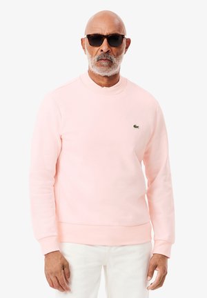 Sudadera de color rosa claro con cuello redondo, mangas largas, puños acanalados y un pequeño logo verde en el pecho. Textura de tela suave.