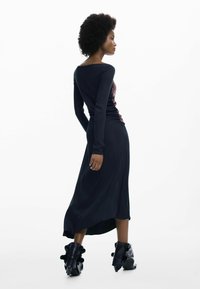 Fekete hosszú ujjú ruha, szűkített felsőrésszel és libegő szoknyával. Oldalán redőzött részletekkel és egyedi fekete fodros csizmákkal.