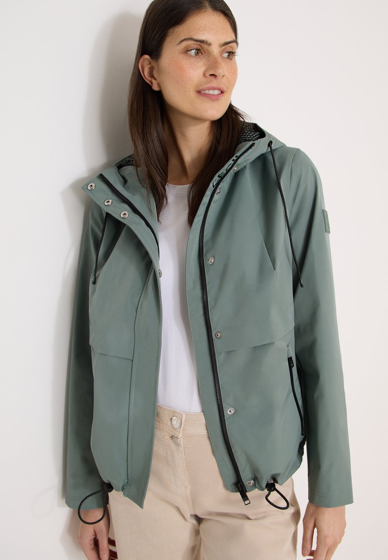 Femme portant une veste à capuche vert sauge par-dessus une chemise blanche et un pantalon beige, regardant sur le côté devant un fond blanc uni.