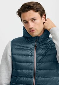 Chaleco acolchado sin mangas con capucha en verde azulado oscuro y diseño acolchado. Cuenta con un cierre frontal completo y mangas de canalé grises.