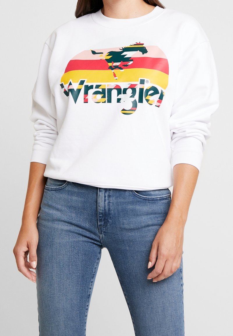 Donna che indossa una camicia a maniche lunghe bianca con un cavallo colorato e il logo "Wrangler", abbinata a jeans in denim blu su uno sfondo chiaro uniforme.