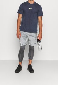 Camiseta texturizada azul marino de Nike con un logo amarillo, pantalones cortos de camuflaje grises, leggings grises y zapatillas negras. Botella de agua clara en la mano.