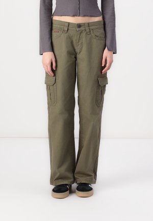 Persoon die olijfgroene cargo-broek met zijzakken draagt, een kort grijs shirt met lange mouwen en zwarte sneakers met beige zolen.