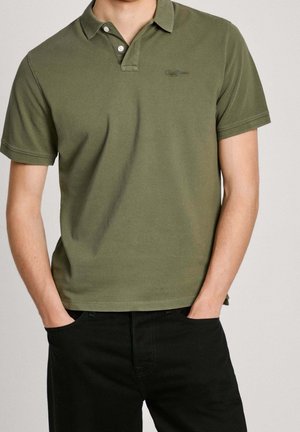 Mężczyzna ubrany w oliwkowo-zieloną koszulkę polo z krótkim rękawem z dwoma białymi guzikami oraz czarne spodnie, stoi z rękami w kieszeniach.